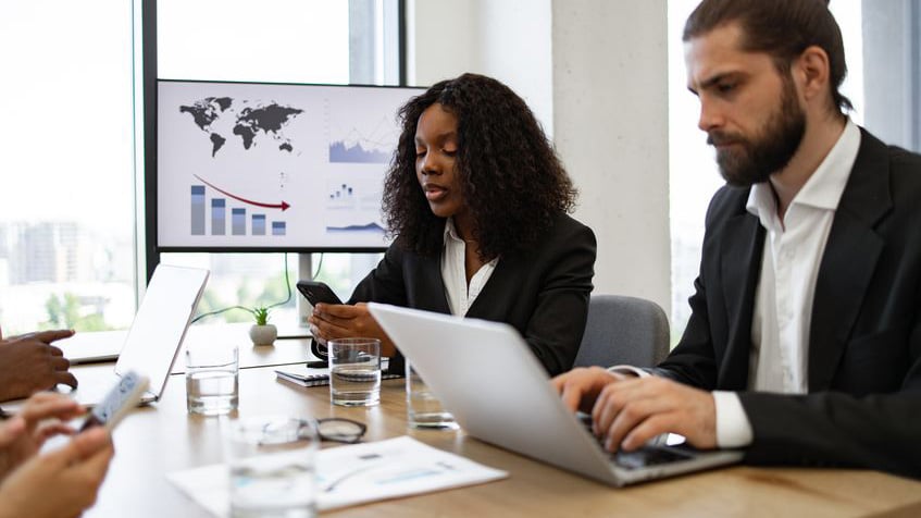 IT professionals sitting at a conference table in a high-pressure war room while a background monitor displays a severe performance decline, representing the manual recovery friction and costly downtime risks that Automic Automation for VMware Cloud Foundation resolves through automated business continuity.